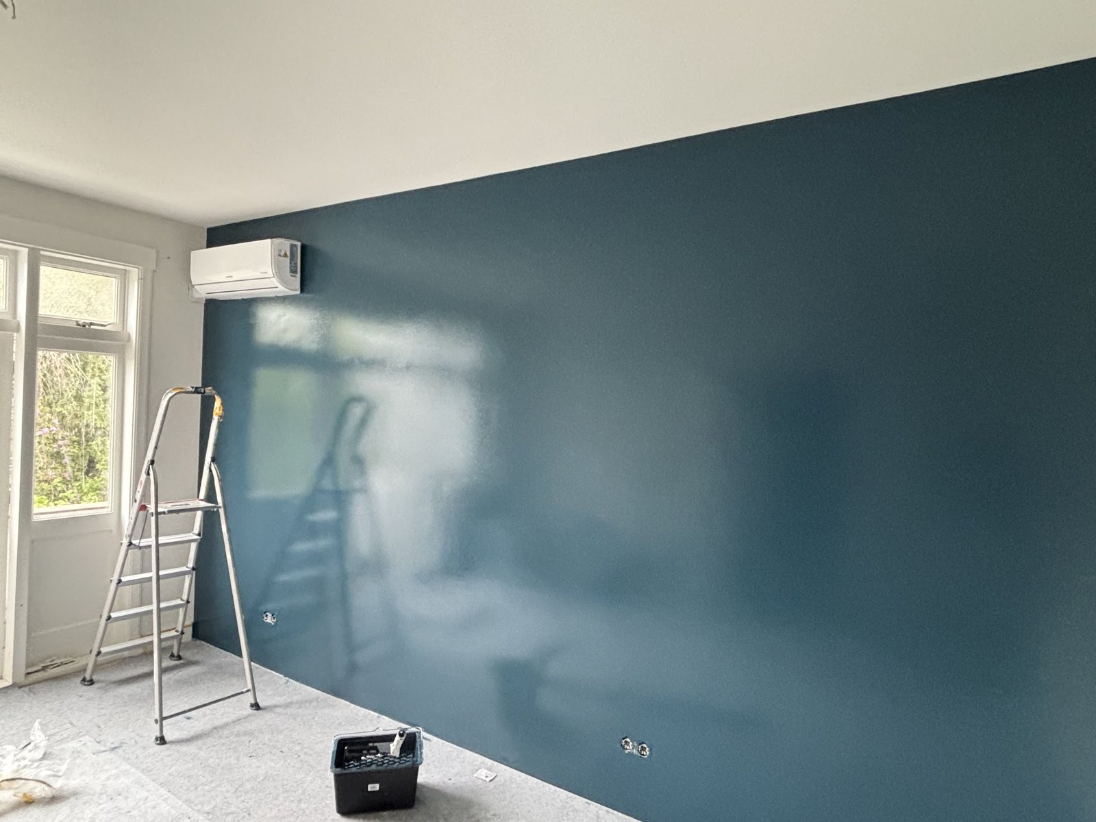Een diep blauwgroene wand in een lege kamer, met een trap en verfbak