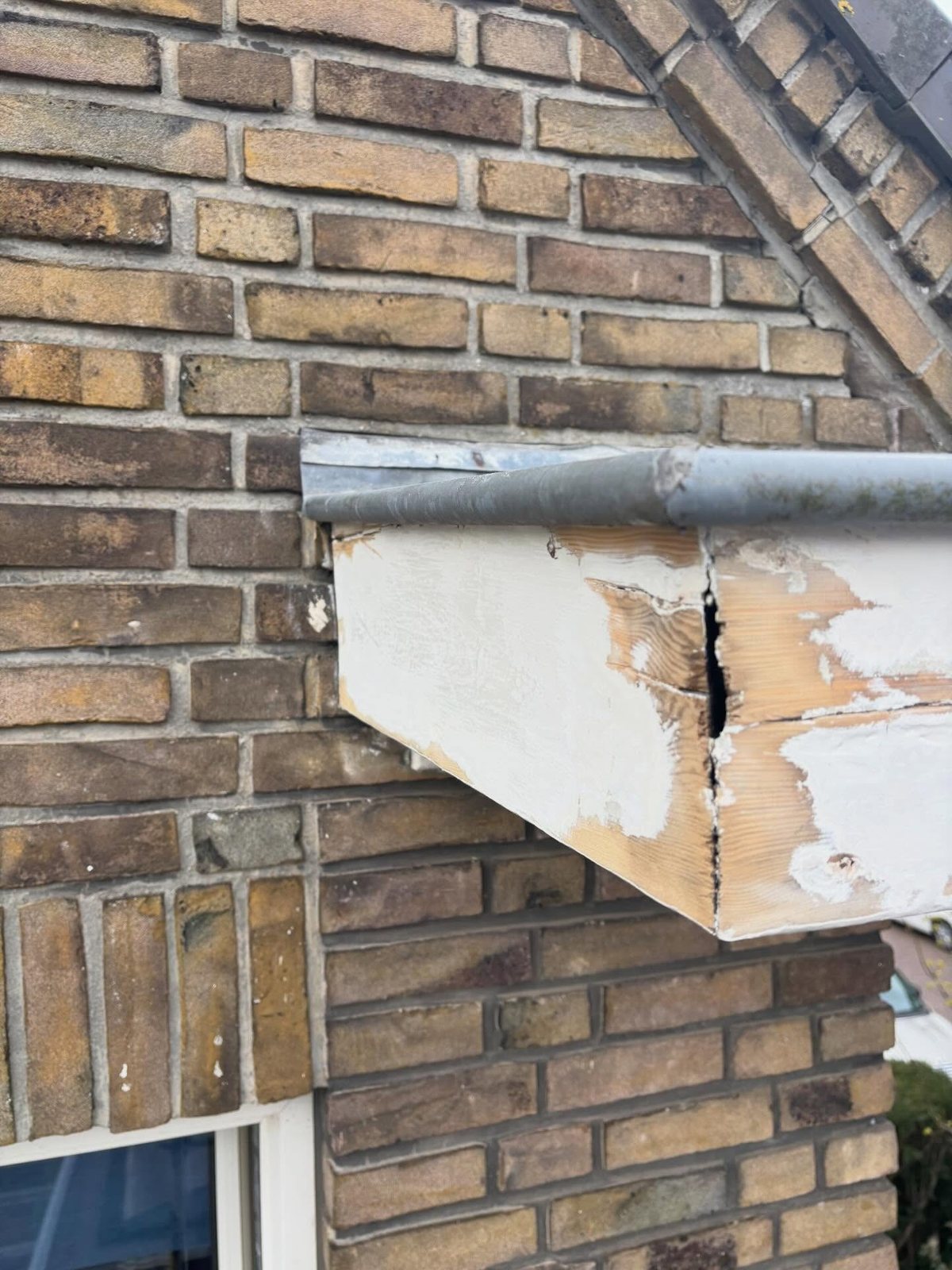 Een aangetaste, afbladderende boeiboord onder een dakrand van een bakstenen gevel